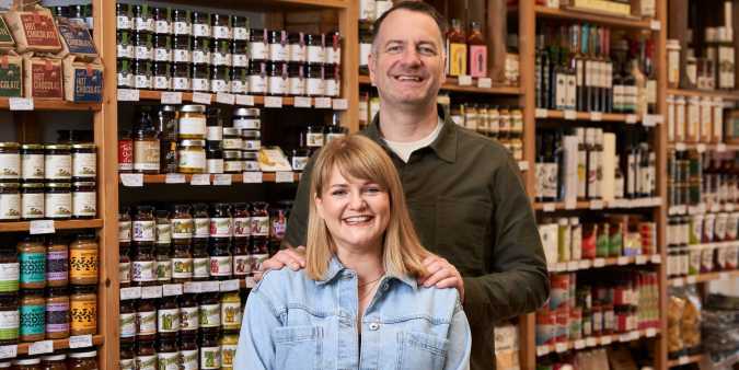 A couple standing in front of a shelf full of colourful deli items.