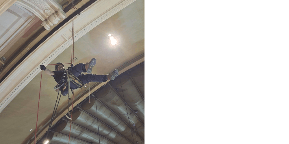 Neill Shimmen wearing a hardhat and safety gloves hanging from a ceiling in a concert hall in a belay.